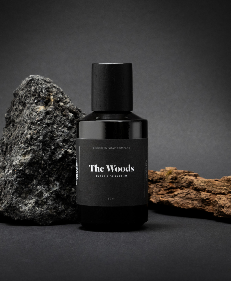 Brooklyn Soap Company The Woods Extrait de Parfum – ein schwarzer Flakon mit minimalistischer Beschriftung, inszeniert vor einem rauen Stein und einem Stück Baumrinde auf dunklem Hintergrund.