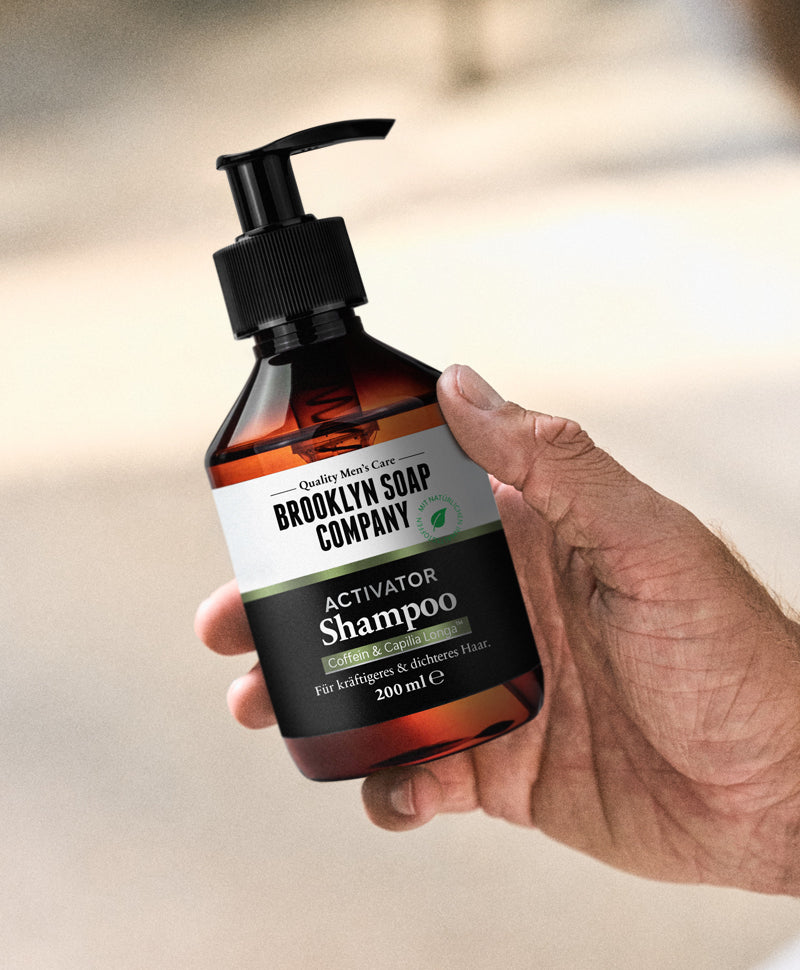 Eine Hand hält eine Flasche Brooklyn Soap Company Activator Shampoo mit Pumpspender. Das Etikett weist auf Inhaltsstoffe wie Coffein & Capilia™ Longa hin sowie auf die Wirkung: für kräftigeres & dichteres Haar.